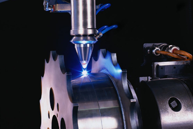 Laserbearbeitungskopf über einer leuchtenden Schweißstelle an einem eingespannten zylindrischen Metallbauteil mit gezahntem Rand