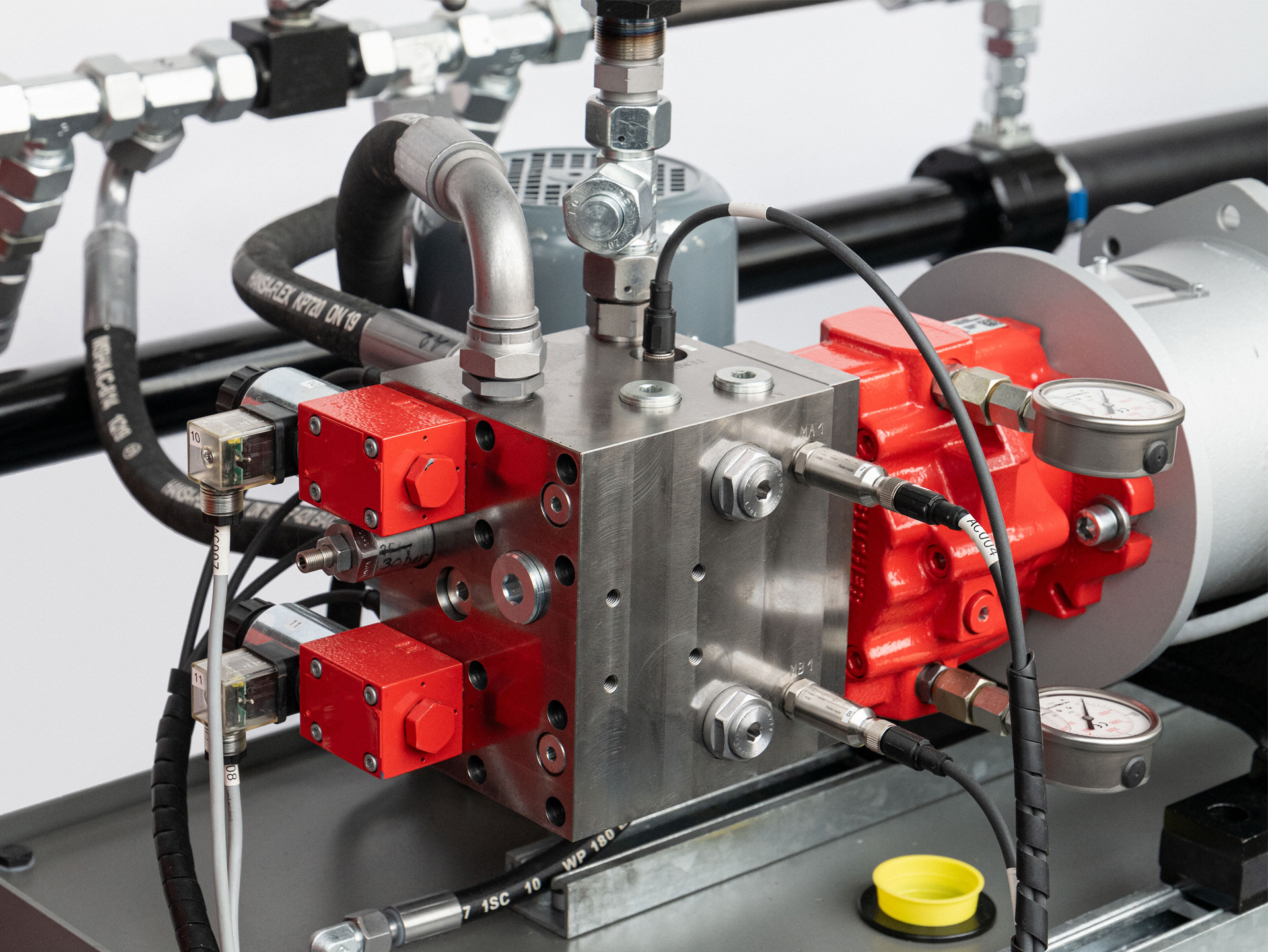 Metallischer Hydraulikblock mit roten Ventilmodulen, Schläuchen, Sensoren, Manometern und angeflanschtem Antrieb.