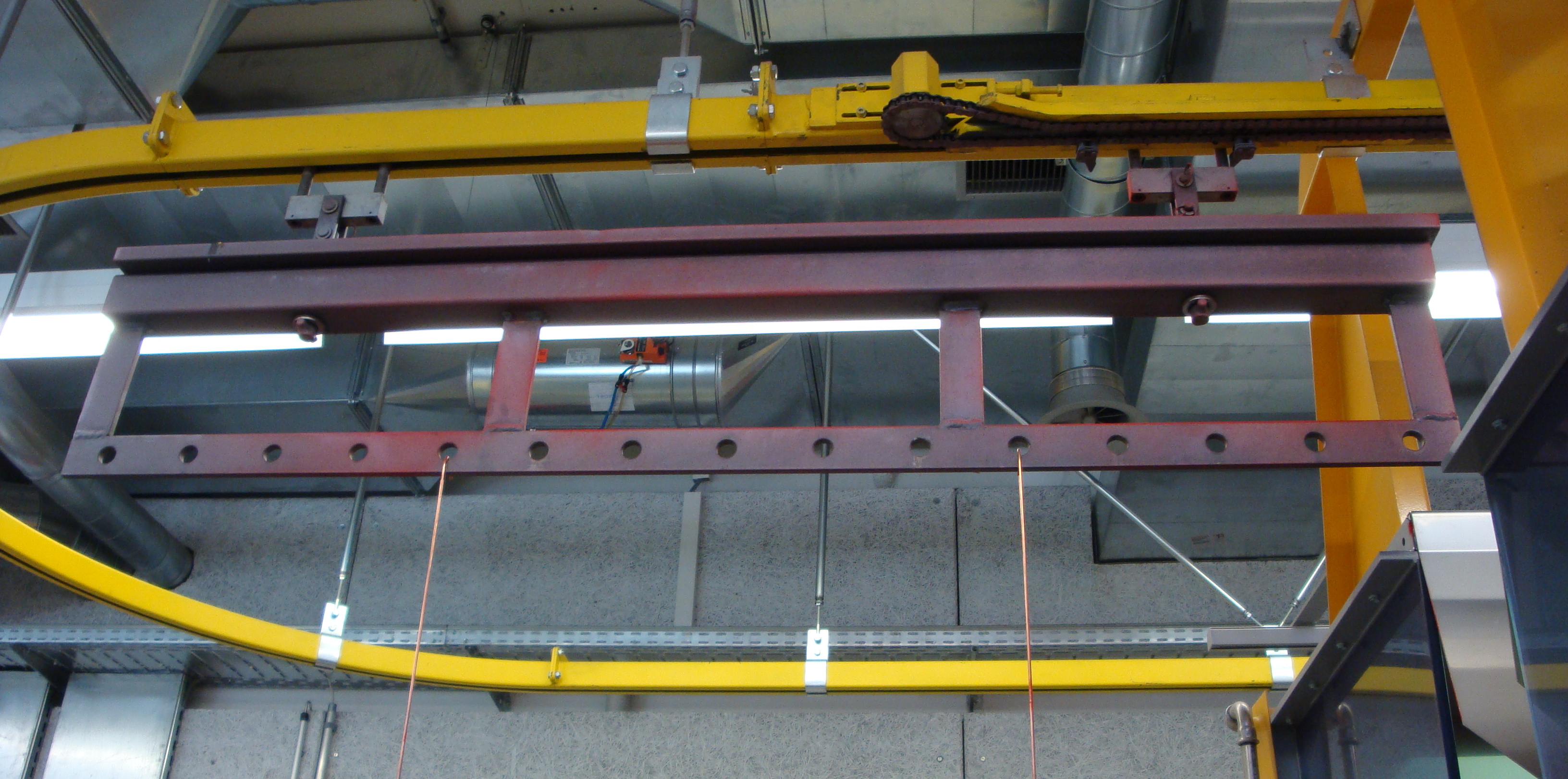Ein Werkstückträger hängt an einer gelben Förderkette in einer Industriehalle.