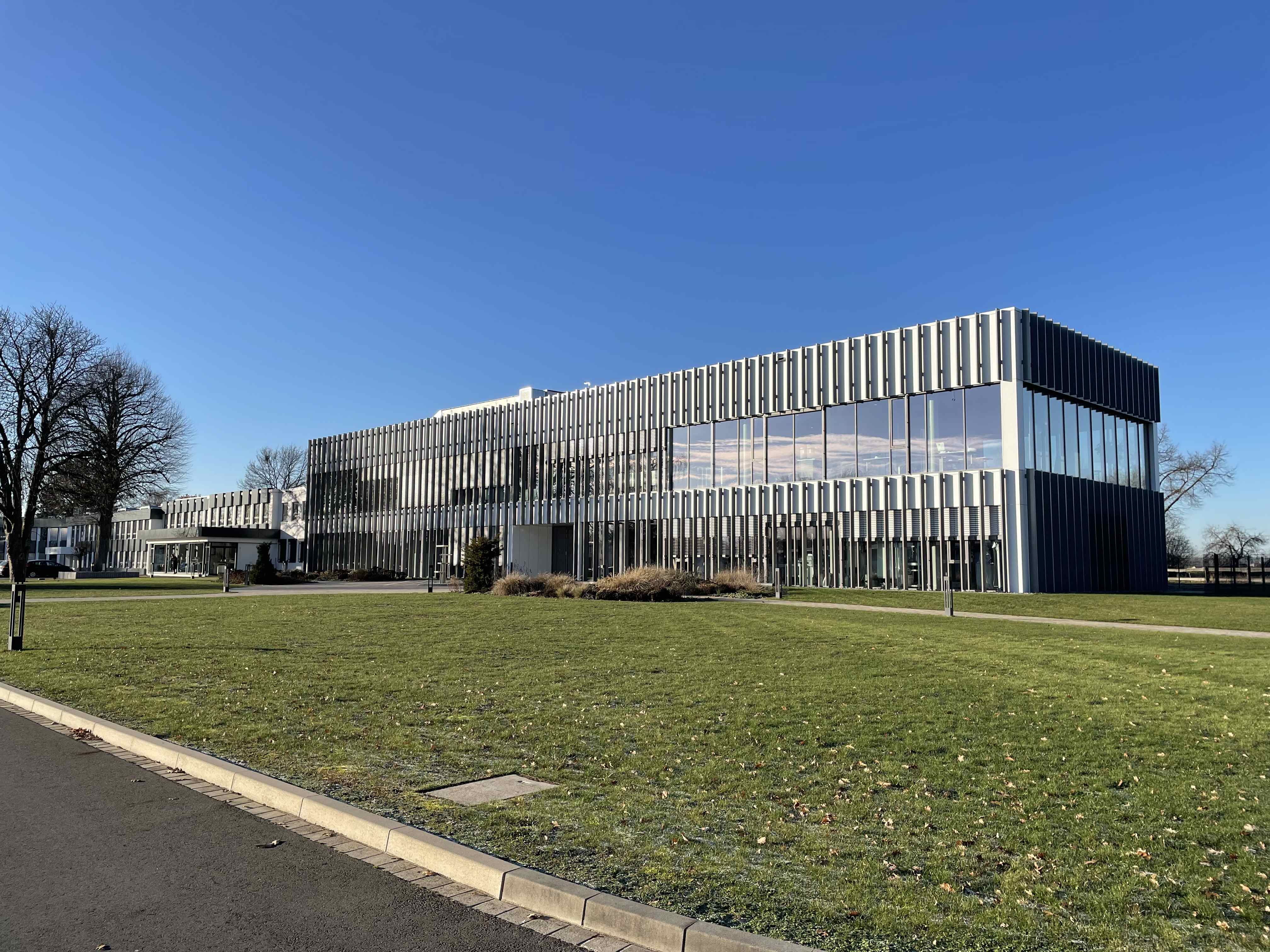 Verglaster zweigeschossiger Gebäudeflügel mit vertikalen Lamellen vor Wiese und blauem Himmel.
