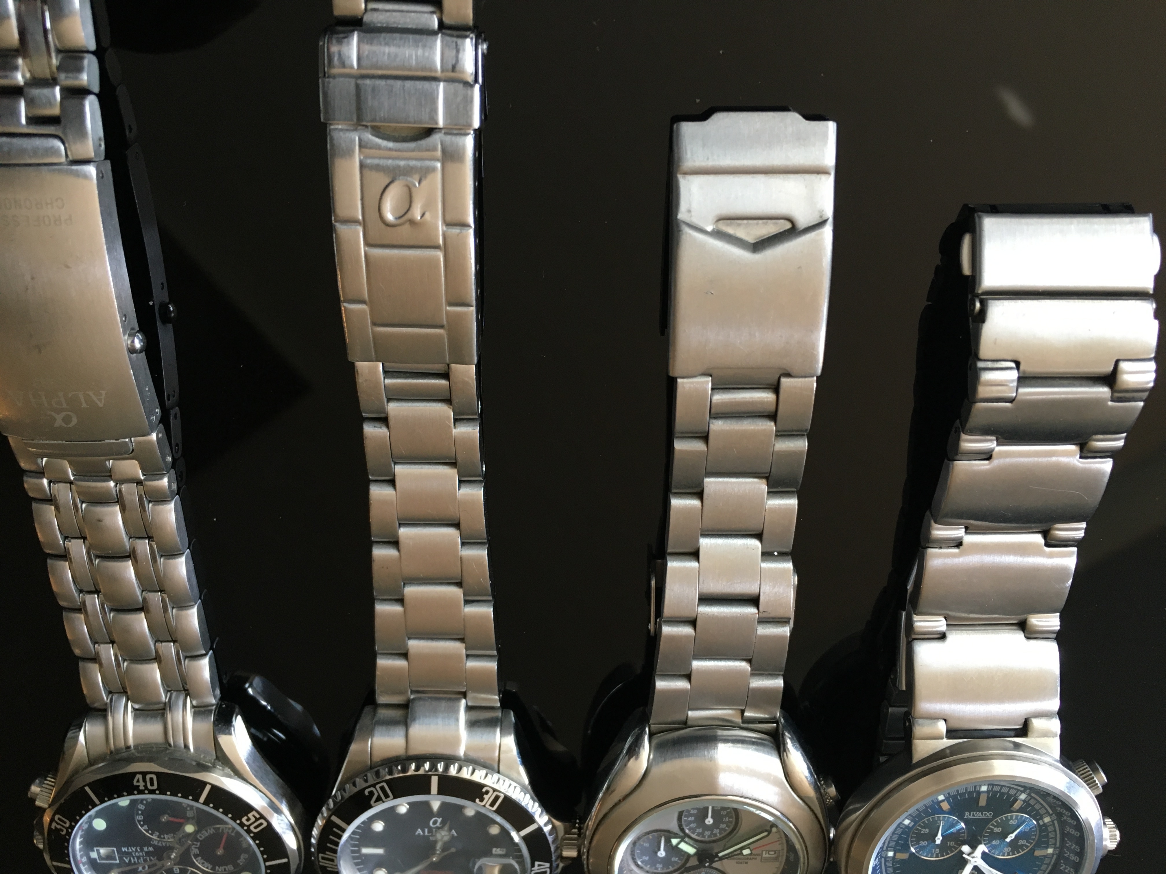 Vier analoge Armbanduhren mit Metallgehäusen und Edelstahlarmbändern auf spiegelnder Unterlage.