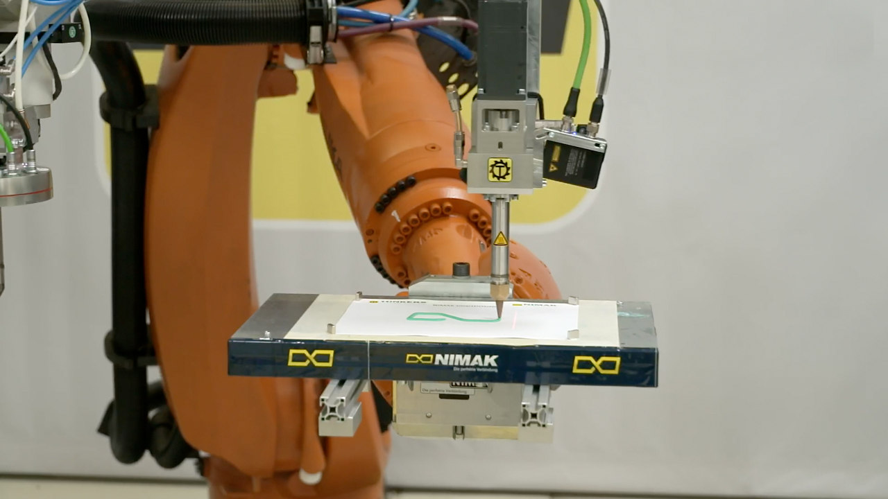 Ein orangefarbener Roboter führt einen schlanken Auftragskopf über eine helle Platte mit grüner Klebstoffraupe.